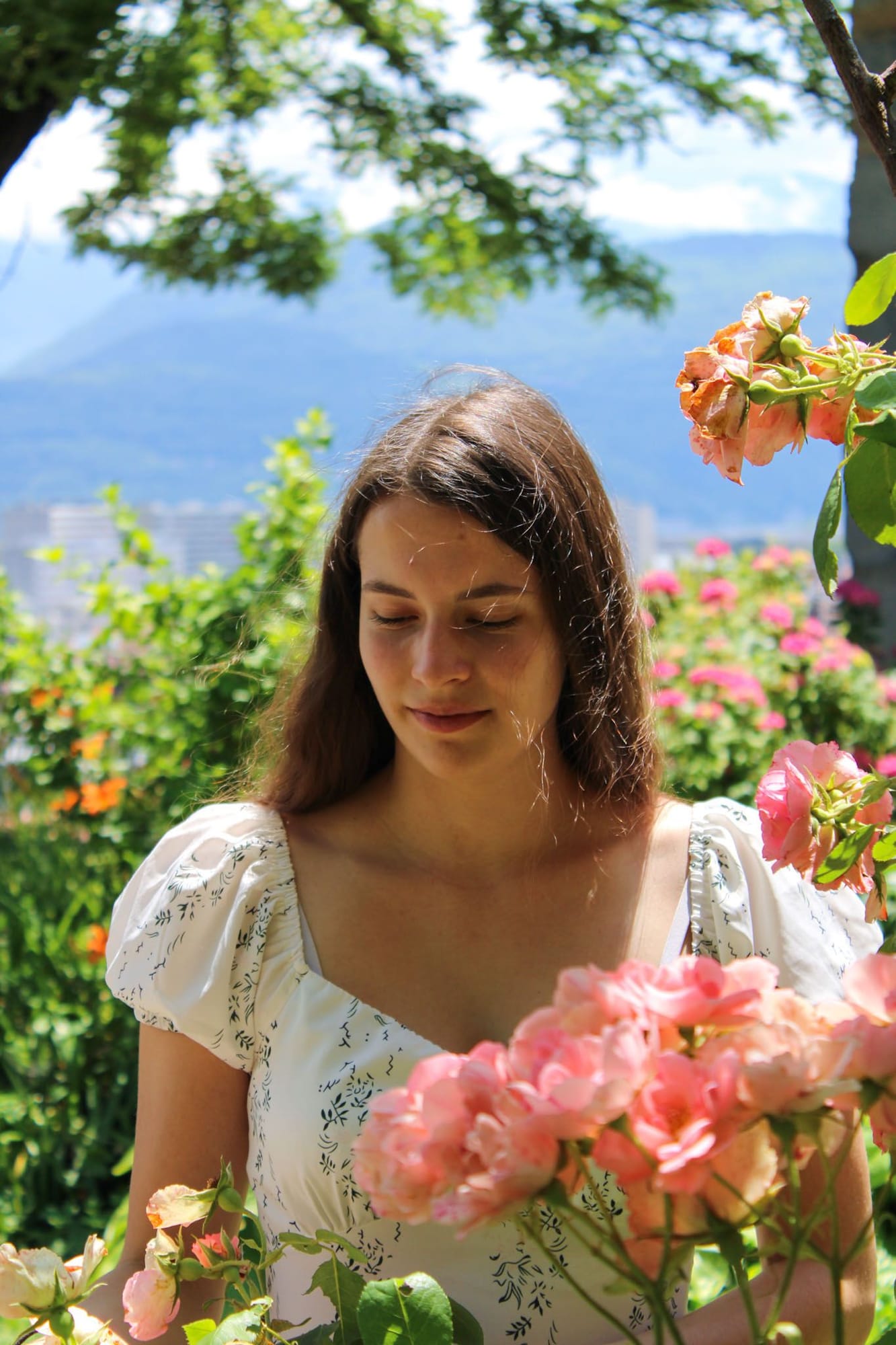 Jeune femme en robe blanche entourée de roses roses, montagnes de Grenoble en arrière-plan, séance photo mariage en plein air