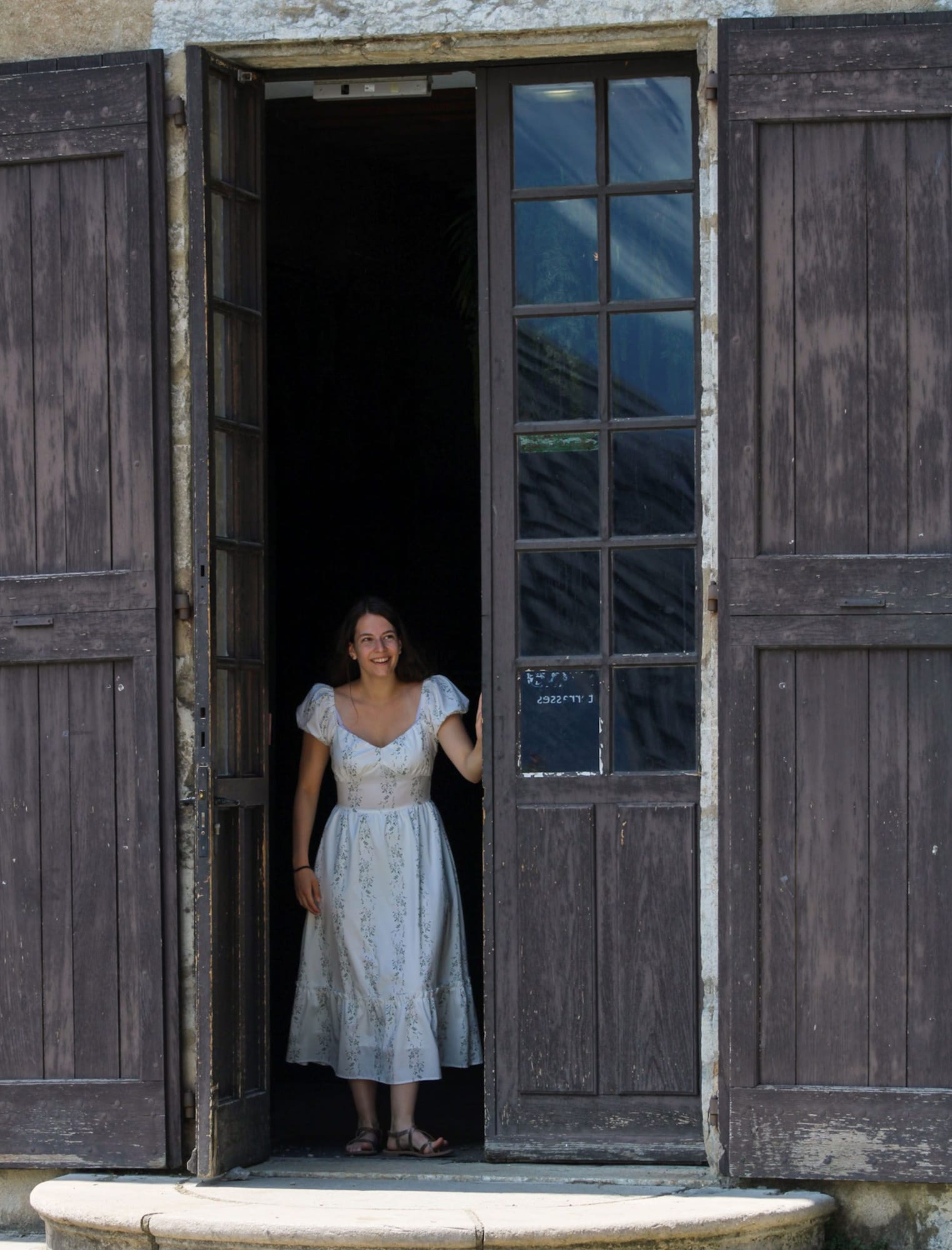 Jeune femme radieuse sur le seuil d'une ancienne porte en bois, robe longue champêtre, inspiration photo de mariage joyeuse à Grenoble